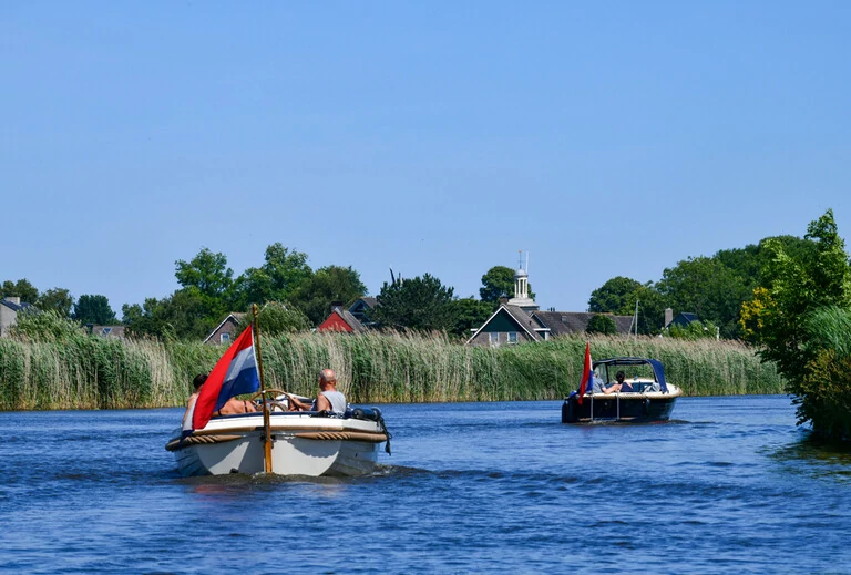 Niedriges Blickwinkel vom Wasser auf zwei Sportmotorboote, die entlang eines Kanals mit Schilfbeeten zwischen den friesischen Seen in den Niederlanden mit einem klaren blauen Himmel fahren;